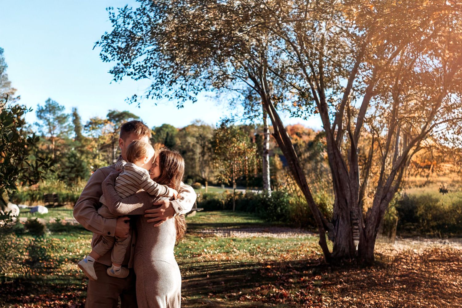 Family of three embracing outdoors in a sunlit autumn park, with a toddler held in a parent's arms beneath a large tree.