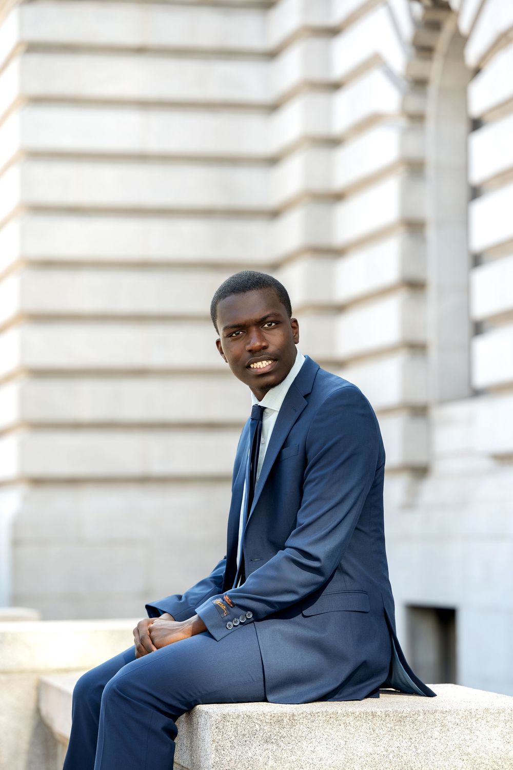 Young Black man in navy suit and tie sitting on stone ledge outside a classic stone building.