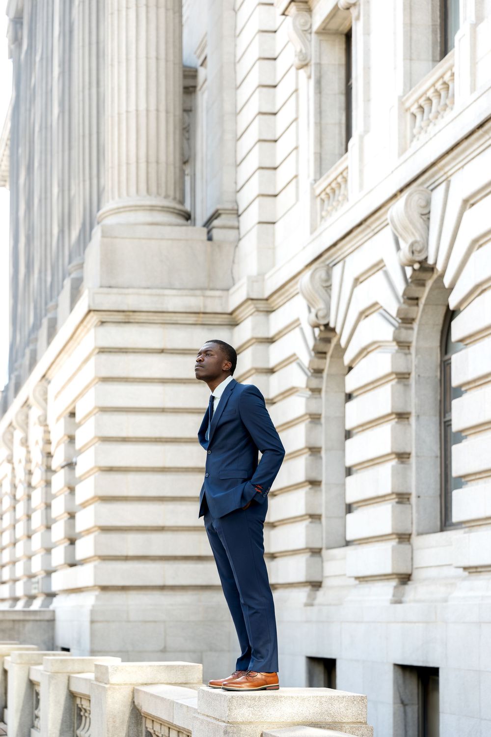 Professional Black businessman in navy suit standing confidently on stone steps of classical architecture building.
