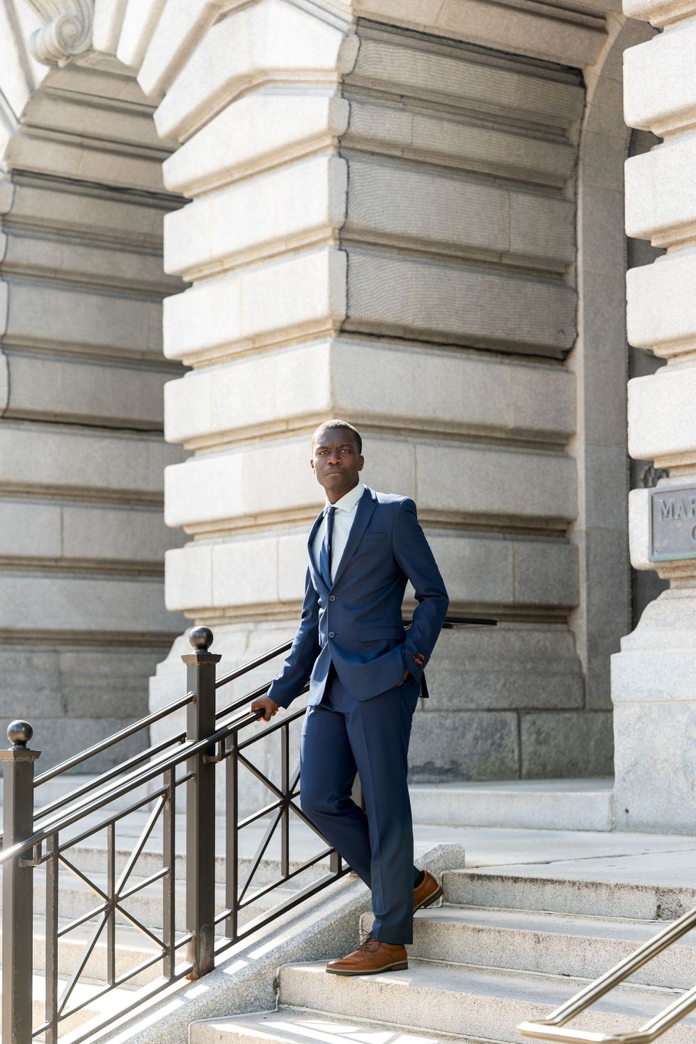 Confident Black man in navy blue suit standing on steps of grand stone building with iron railing.