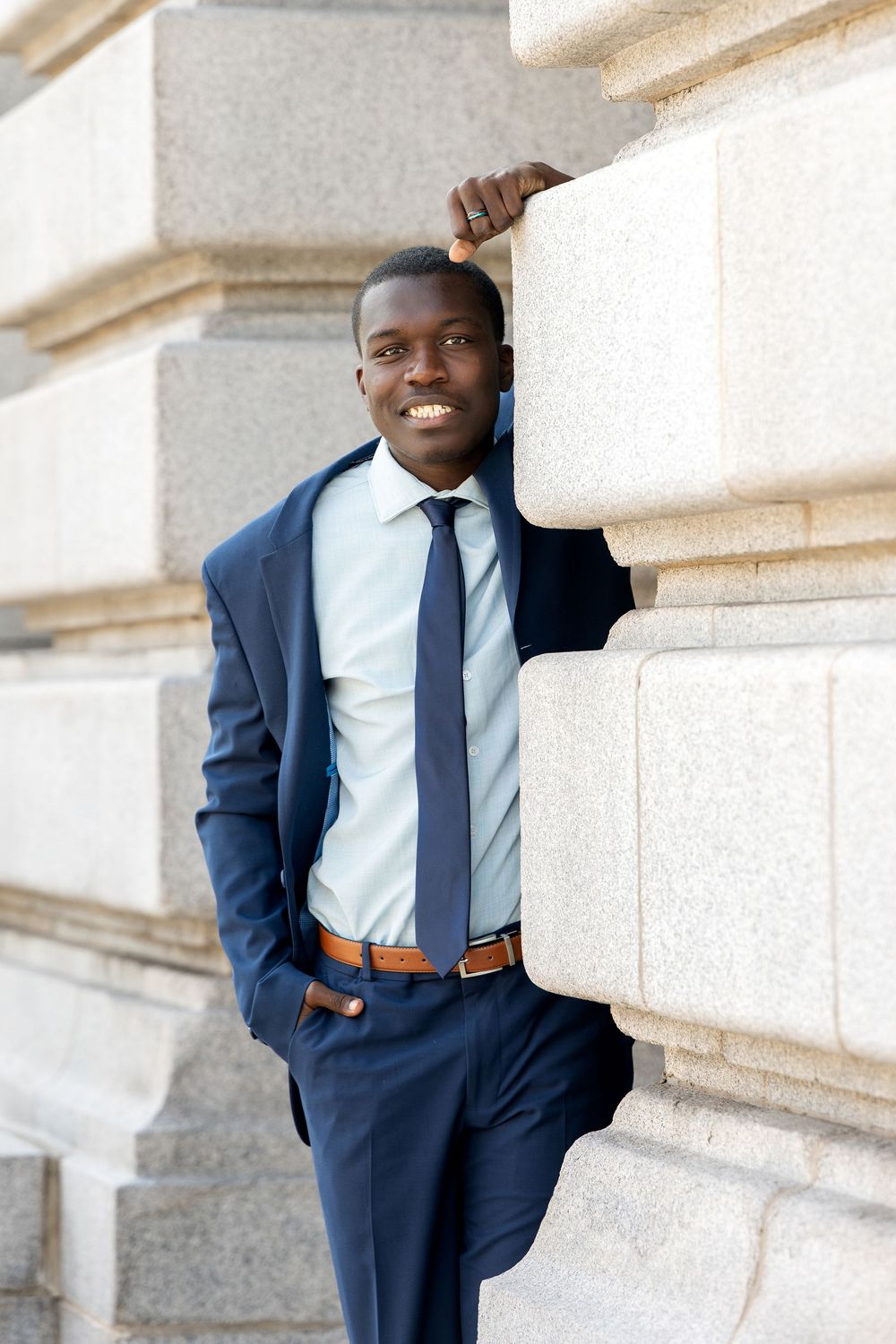 Smiling Black businessman in navy suit and tie leaning against stone columns outdoors.
