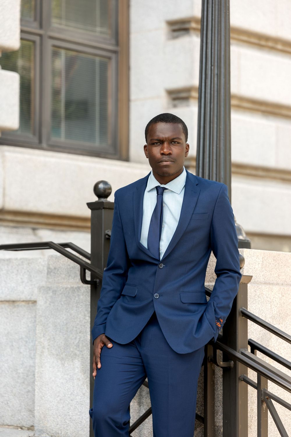 A professional Black man in a navy blue suit and tie stands confidently on outdoor steps near a building.