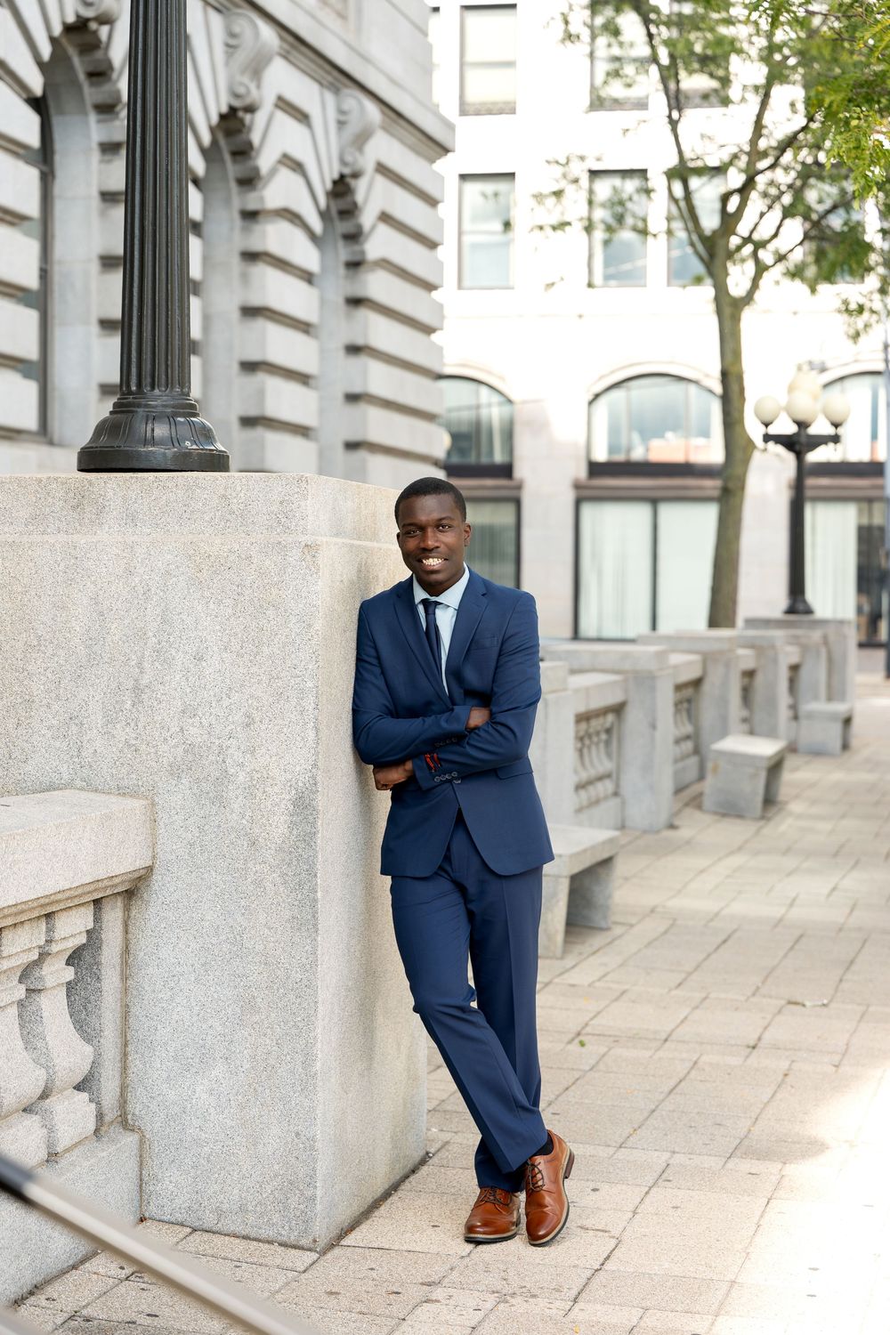 Professional Black businessman in navy suit with crossed arms leaning against stone pillar on urban sidewalk.
