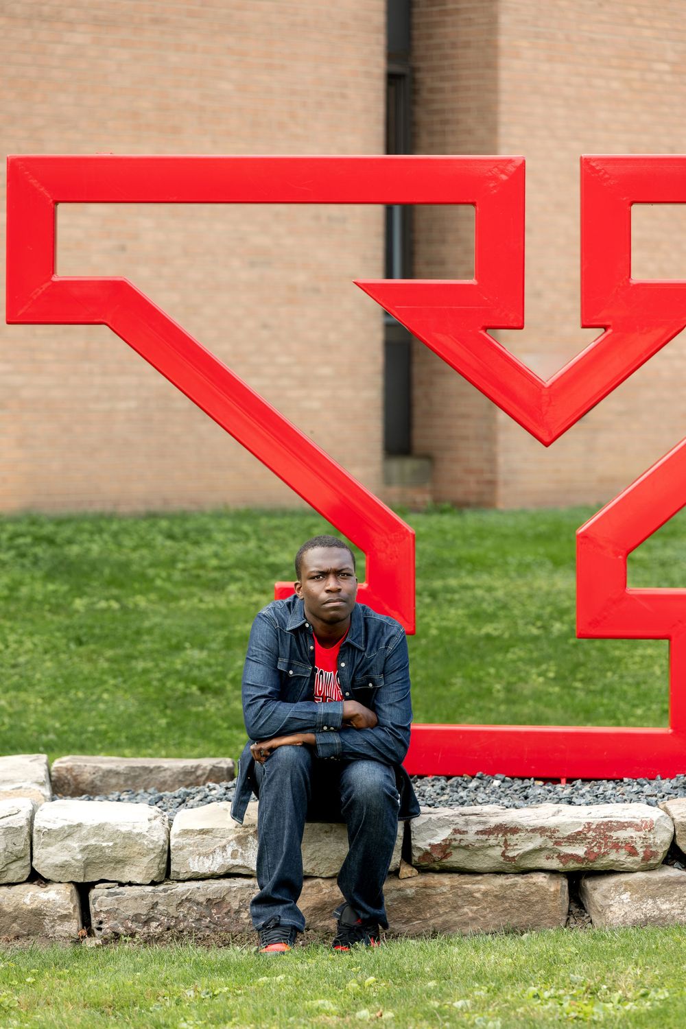 Young Black man in denim jacket sitting on stone ledge near large red sculpture.