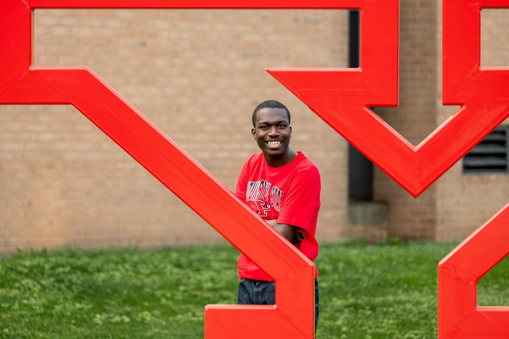 Smiling young Black man in red shirt posing beside large red outdoor sculpture.