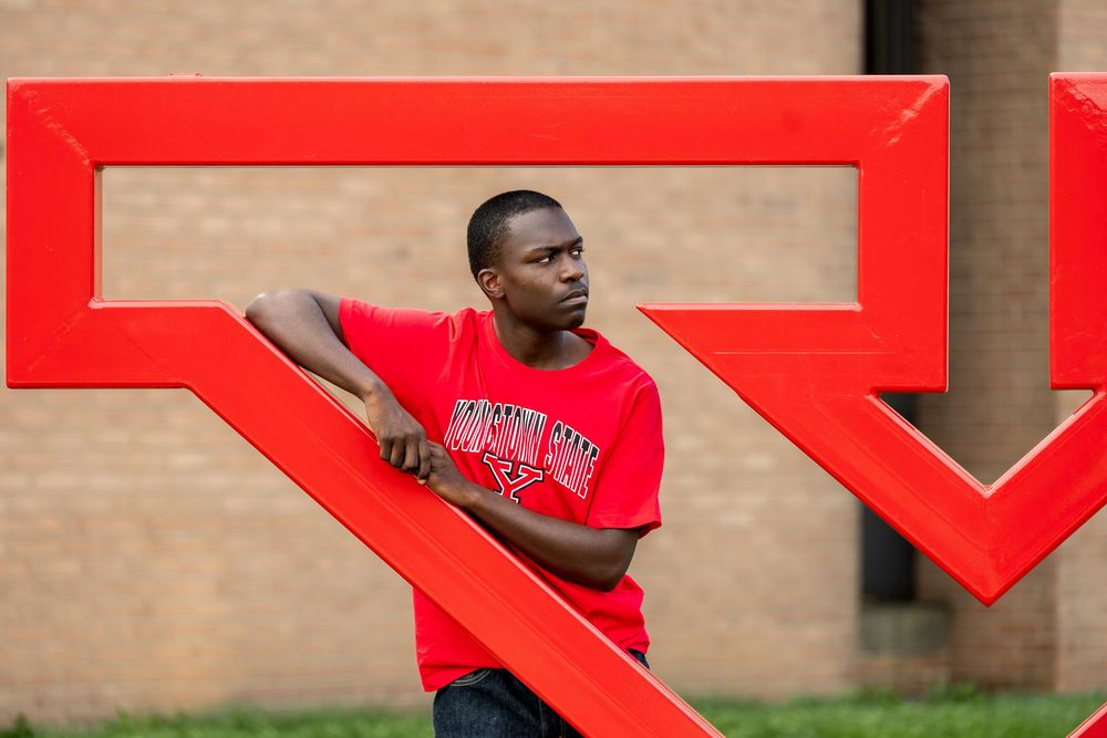 Young Black man in red university t-shirt posing with large red letter sculpture on campus.
