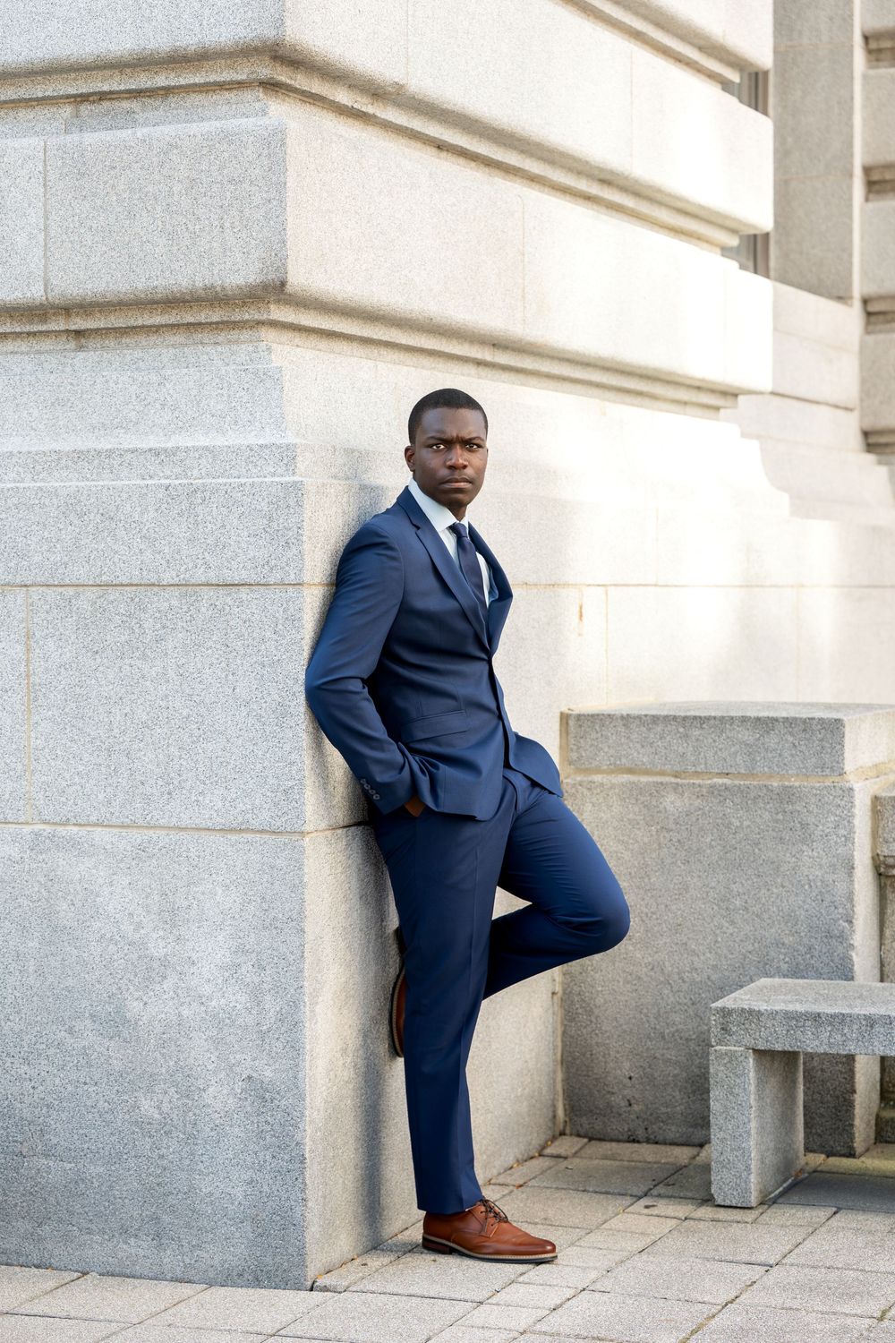 Young Black man in navy blue suit leaning against stone building column outdoors.