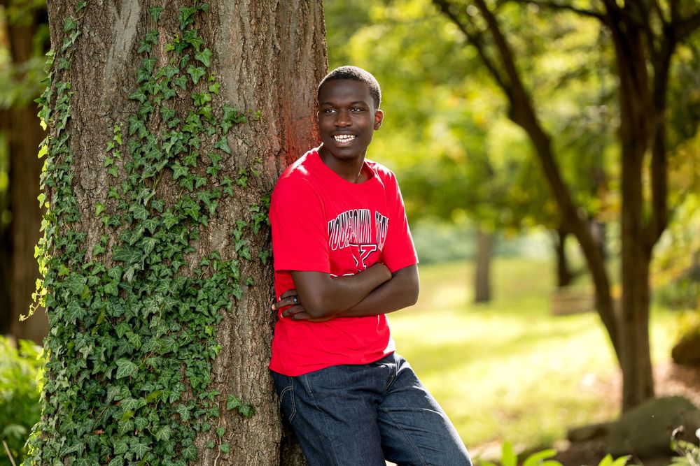 Smiling young Black man in red shirt leaning against ivy-covered tree in park.