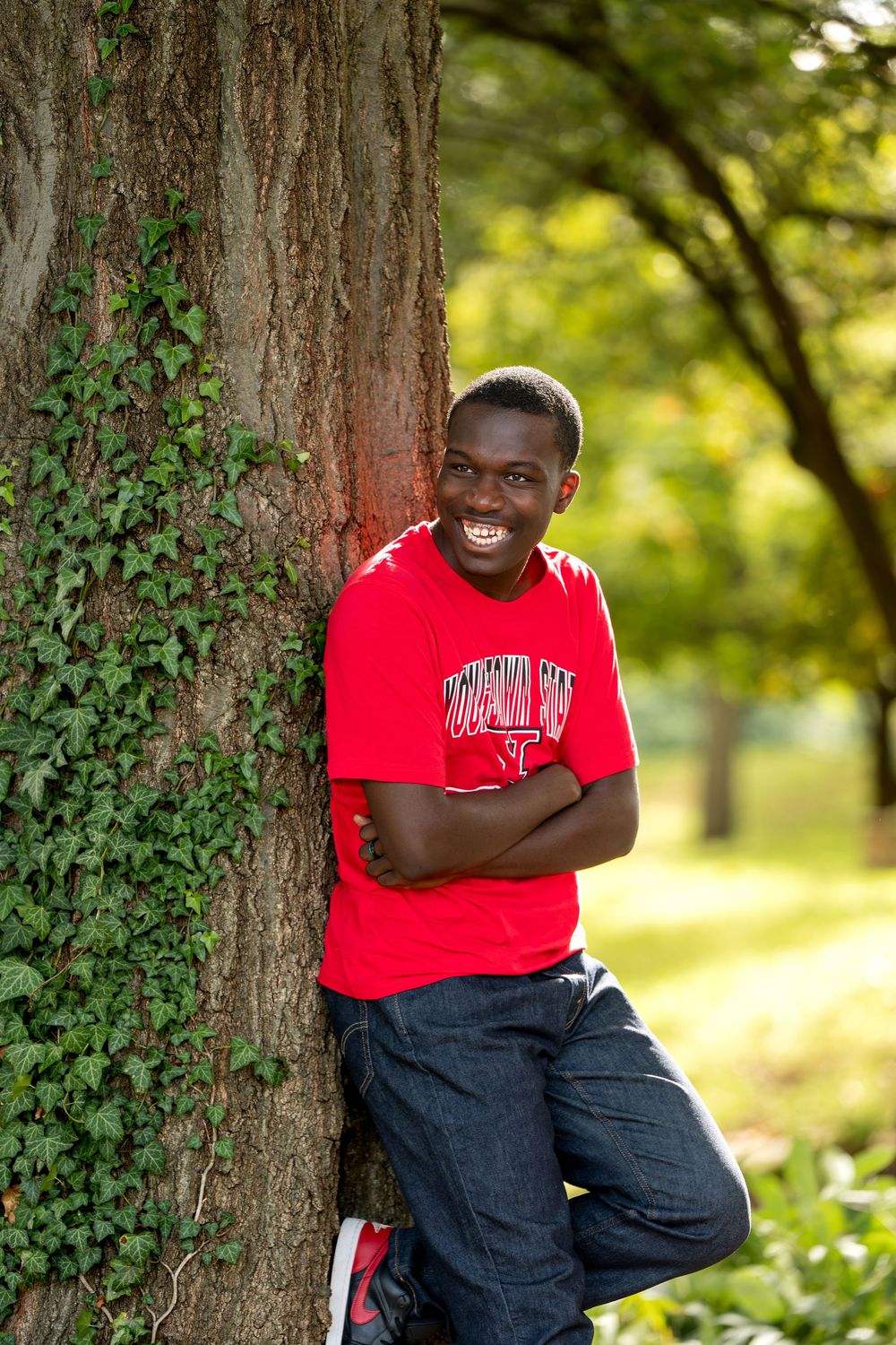 Happy young man in red college shirt leans against ivy-covered tree with arms crossed outdoors.