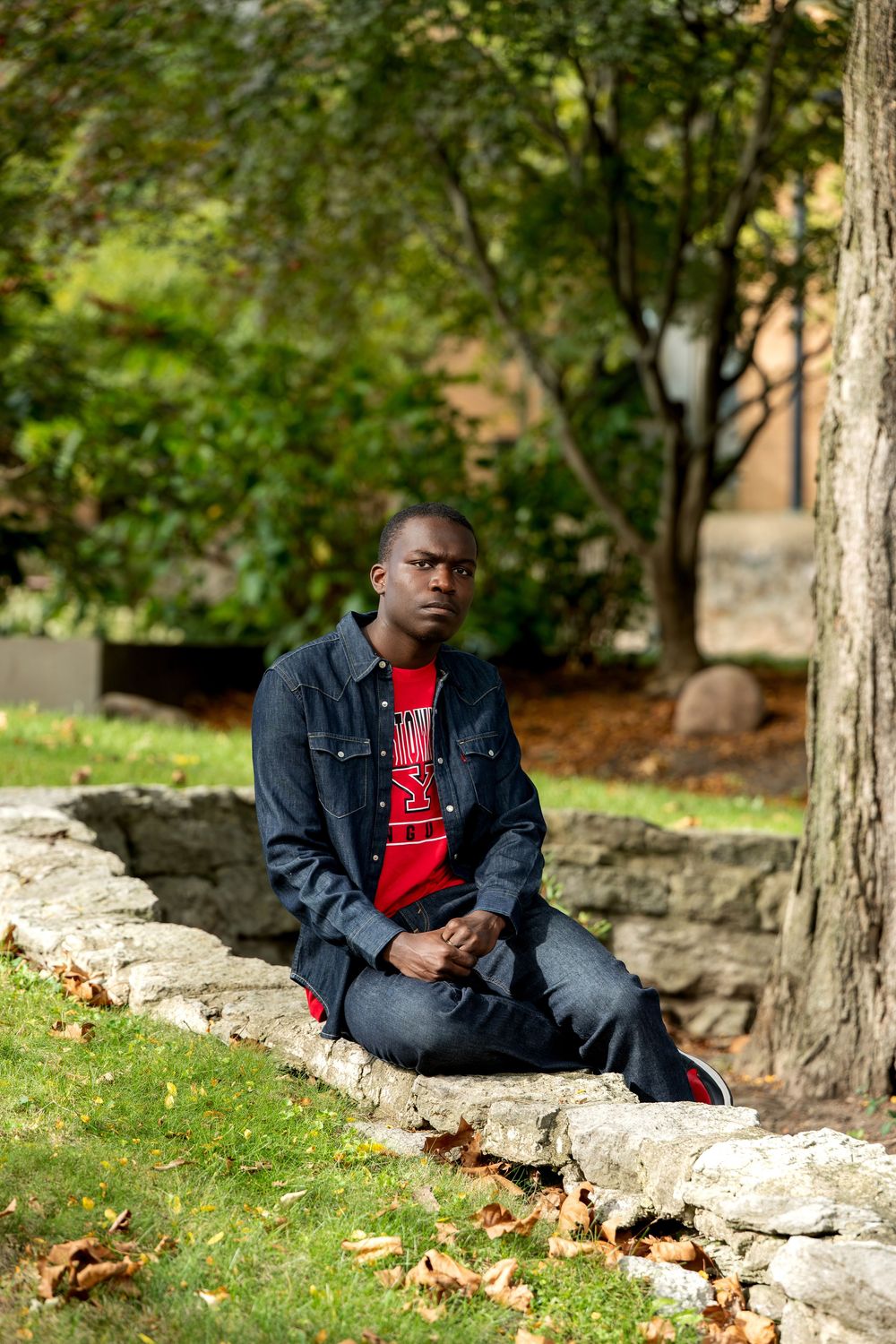 Young Black man in denim jacket and red shirt sitting on stone wall in a park.