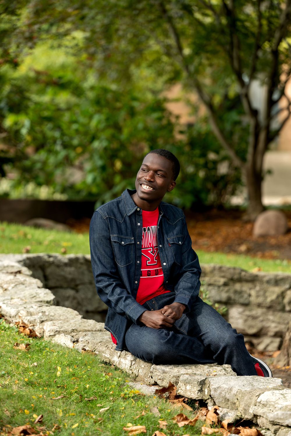 Smiling young Black man in red shirt and denim jacket sitting on stone wall in green park, laughing joyfully.
