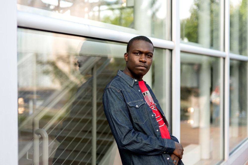 Young man in denim jacket and red tie leaning against modern glass building exterior.
