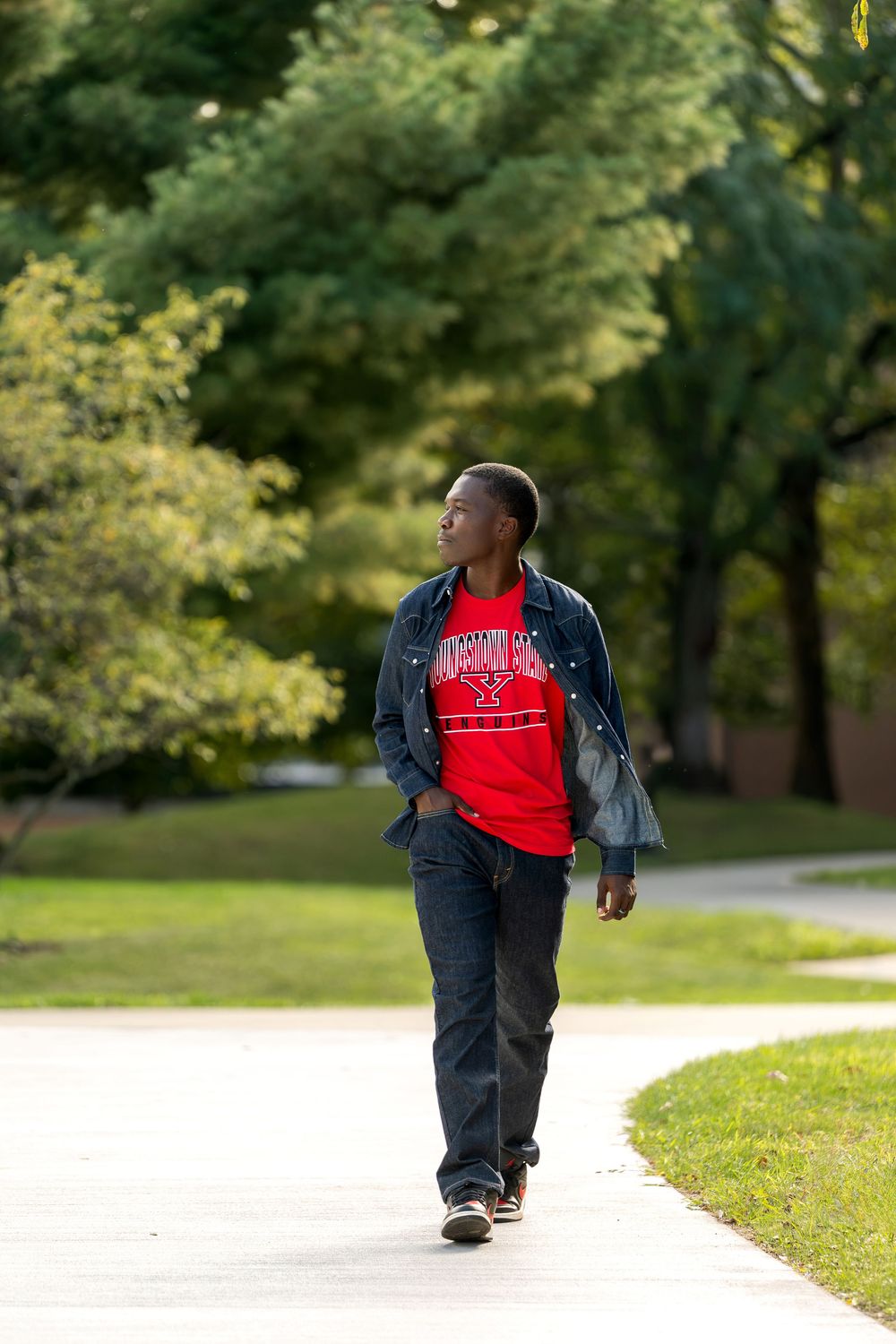 Young Black man strolling on sunny campus walkway in denim jacket and red graphic t-shirt.