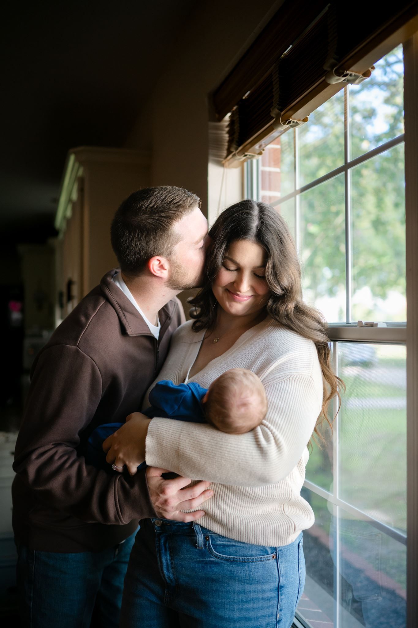 New parents holding their three-week-old baby boy near a window in their home.