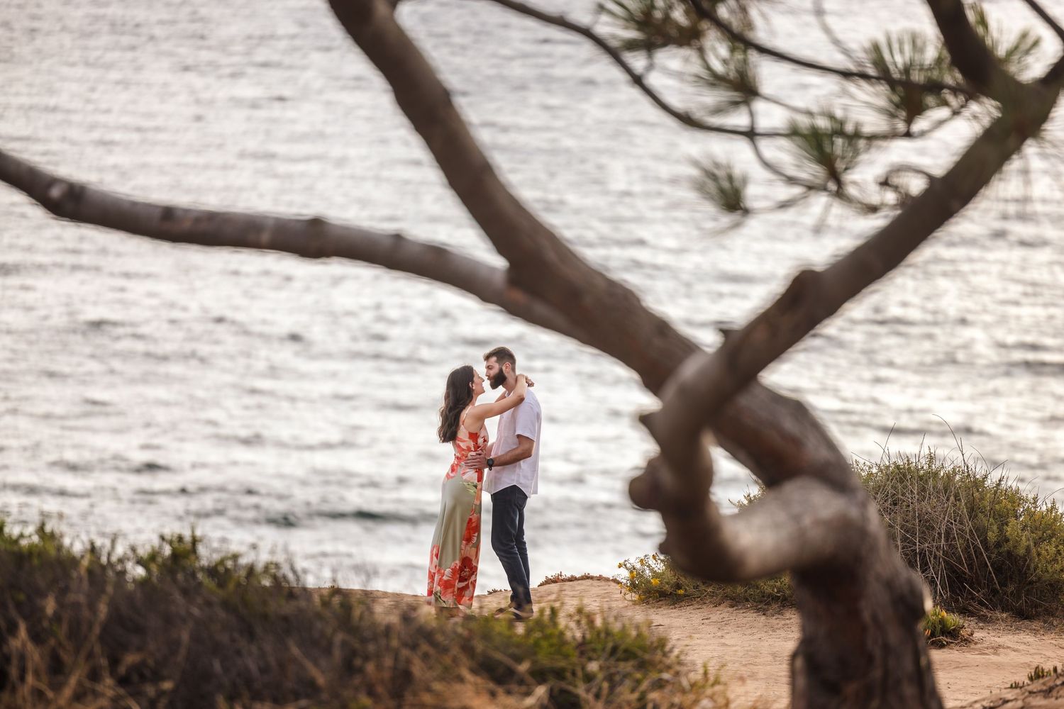 Couple embracing while standing on a coastal cliff overlooking the ocean surrounded by golden wildflowers.