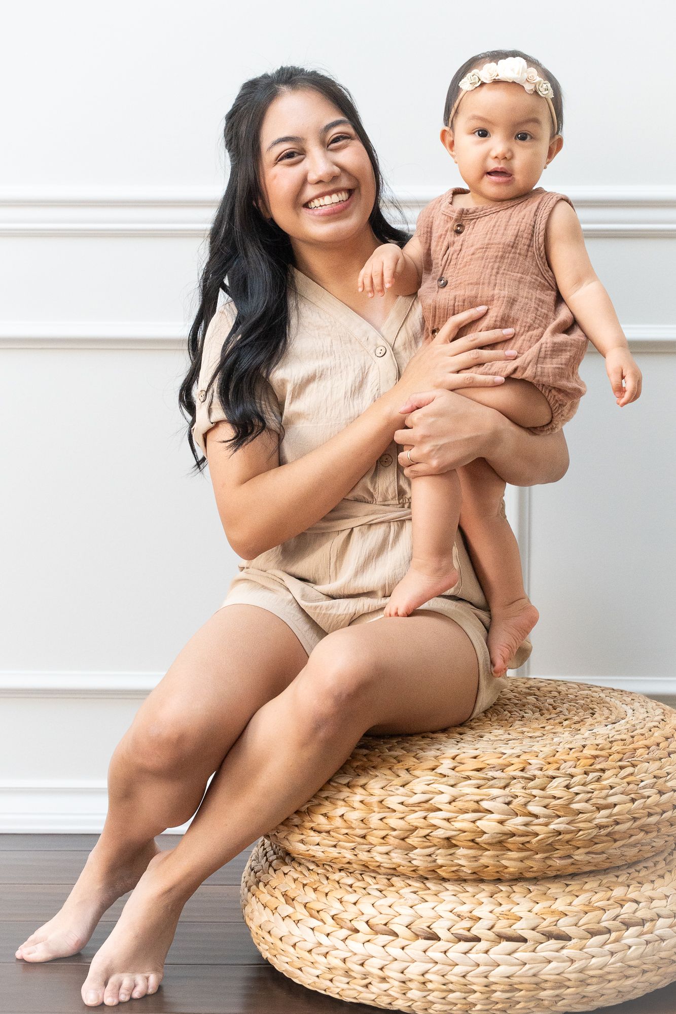 Smiling mother seated on a woven stool, holding her baby in a neutral-toned studio.