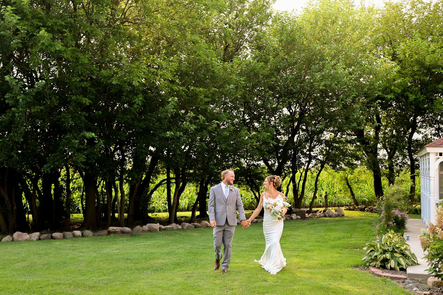 Bride and groom walk hand-in-hand across a lush green lawn surrounded by tall trees on their wedding day.