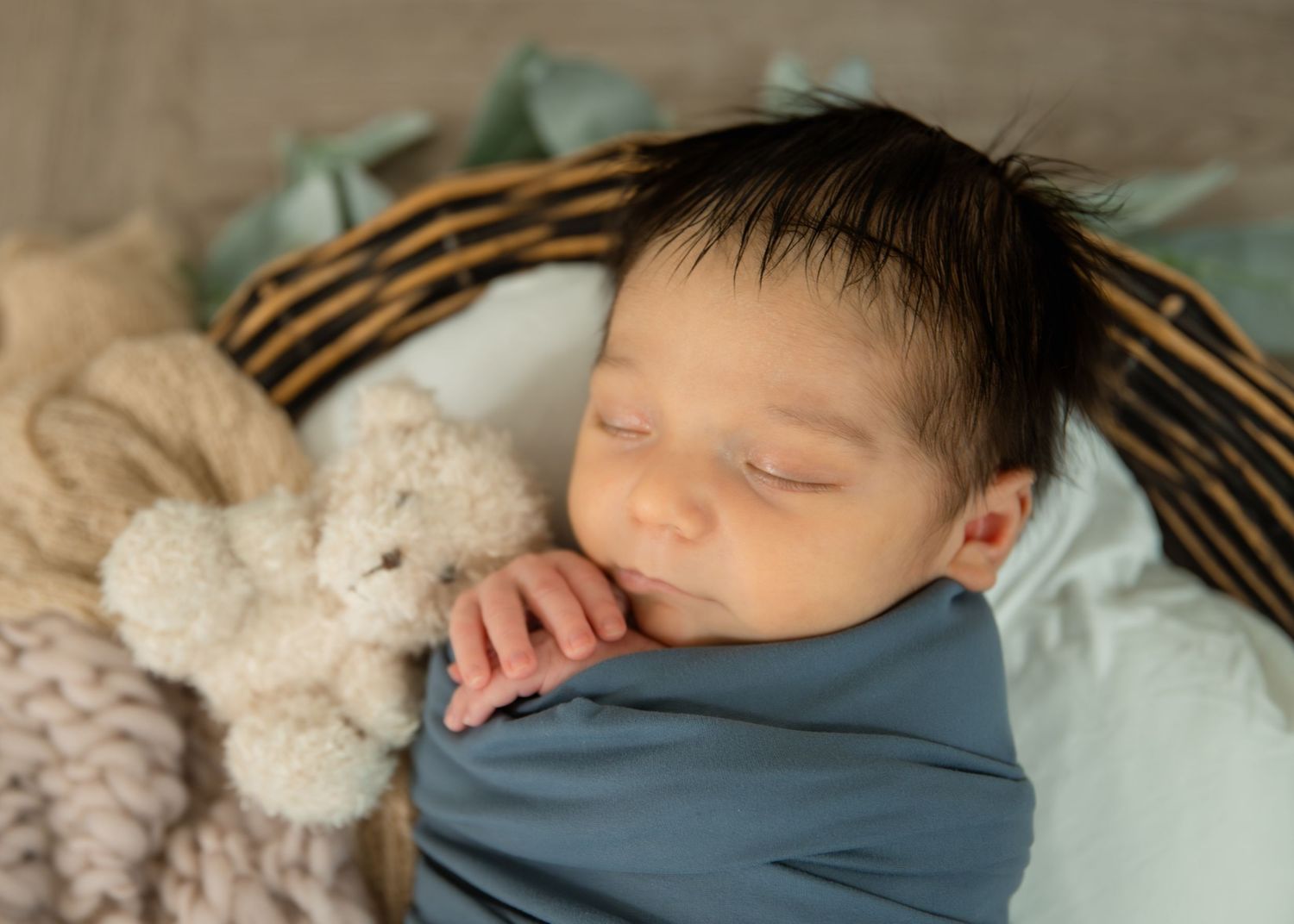 Sleeping newborn baby wrapped in blue swaddle inside a wicker basket with a soft teddy bear.