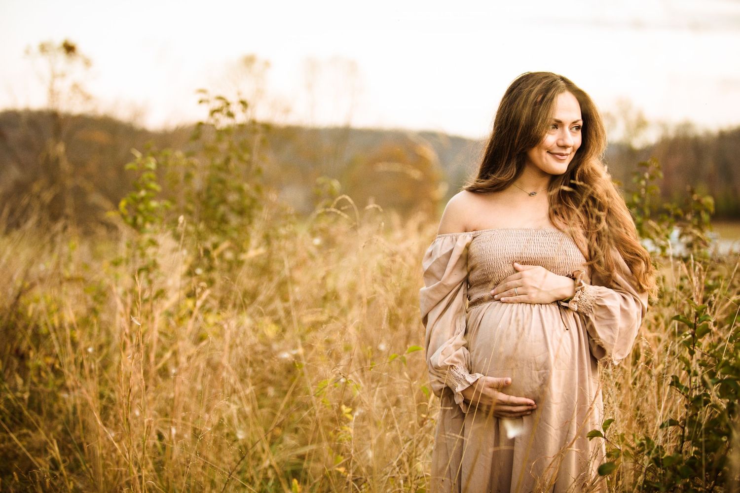 Pregnant woman in beige off-shoulder gown standing in golden field at sunset, cradling baby bump.