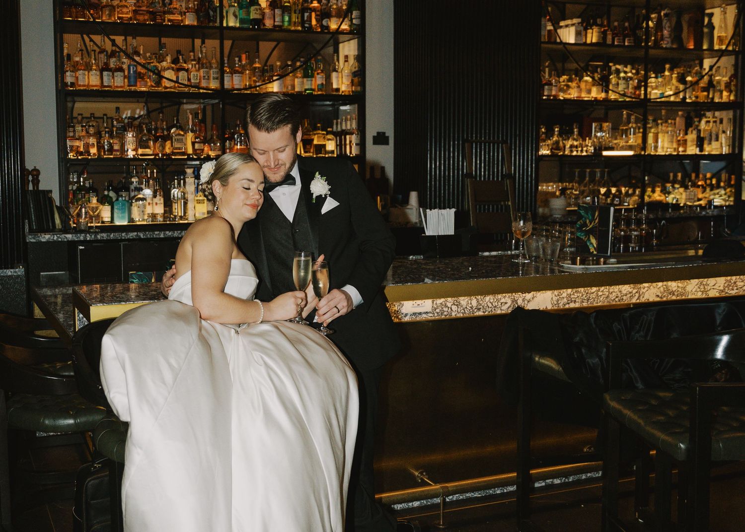 Bride and groom share champagne at an elegant dark bar with backlit bottles during their wedding reception.
