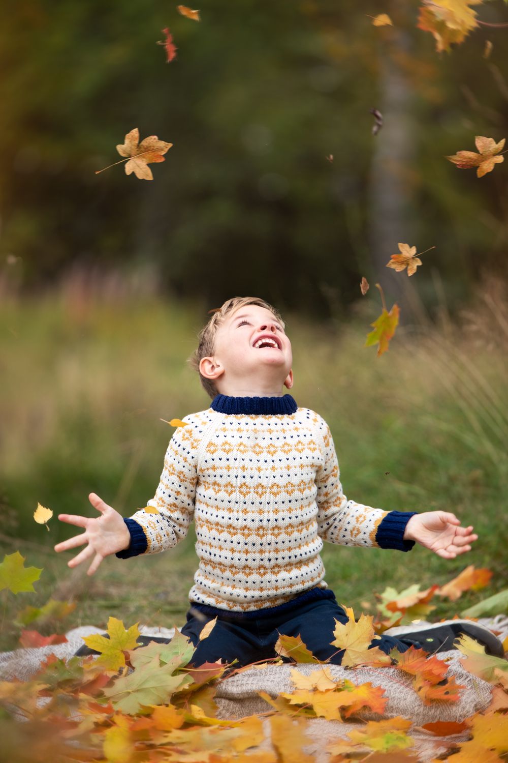 Joyful boy in patterned sweater tossing autumn leaves in the air outdoors, laughing with arms outstretched.