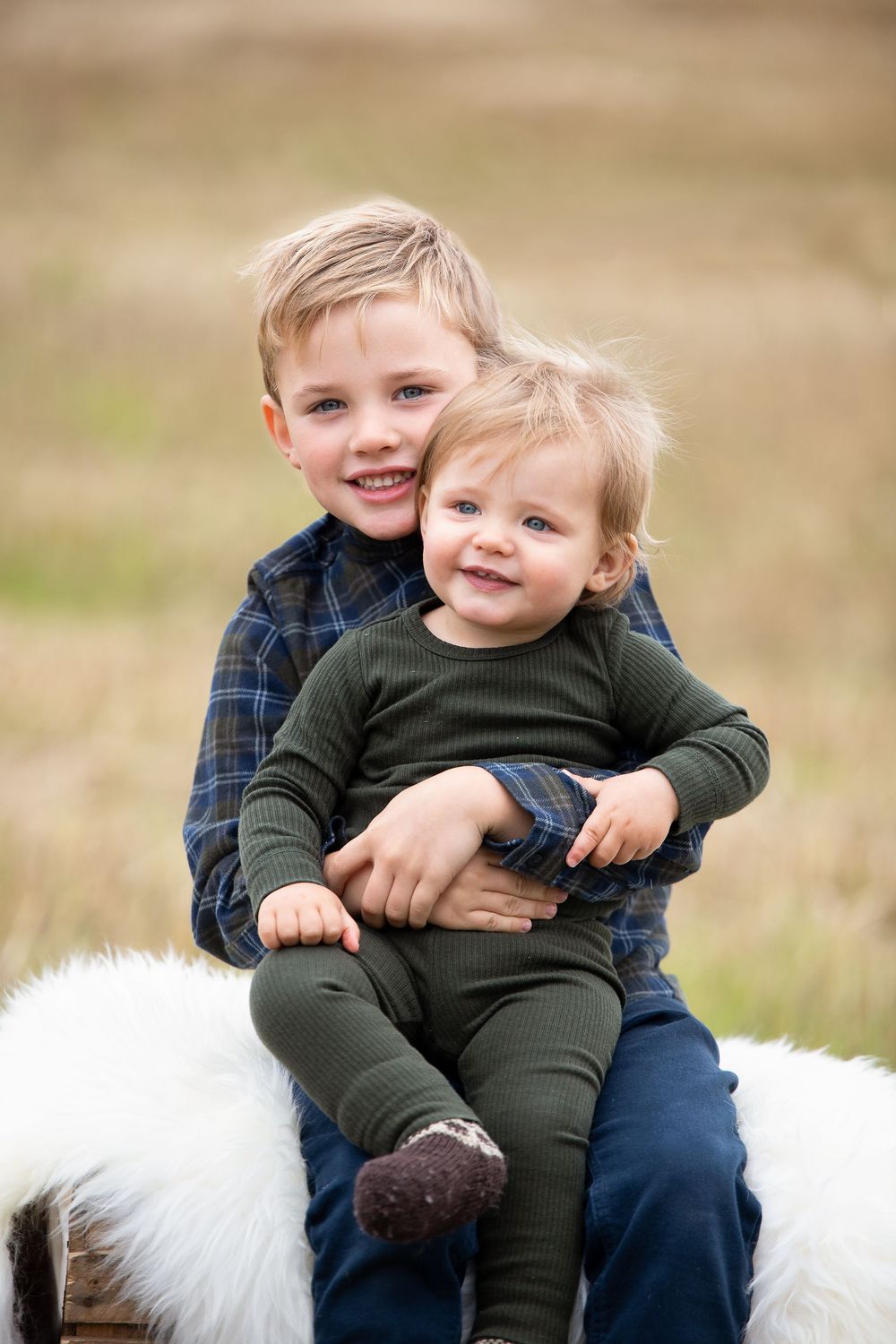 Two young blonde brothers sitting outdoors, older boy hugging toddler sibling, both smiling in coordinating outfits.