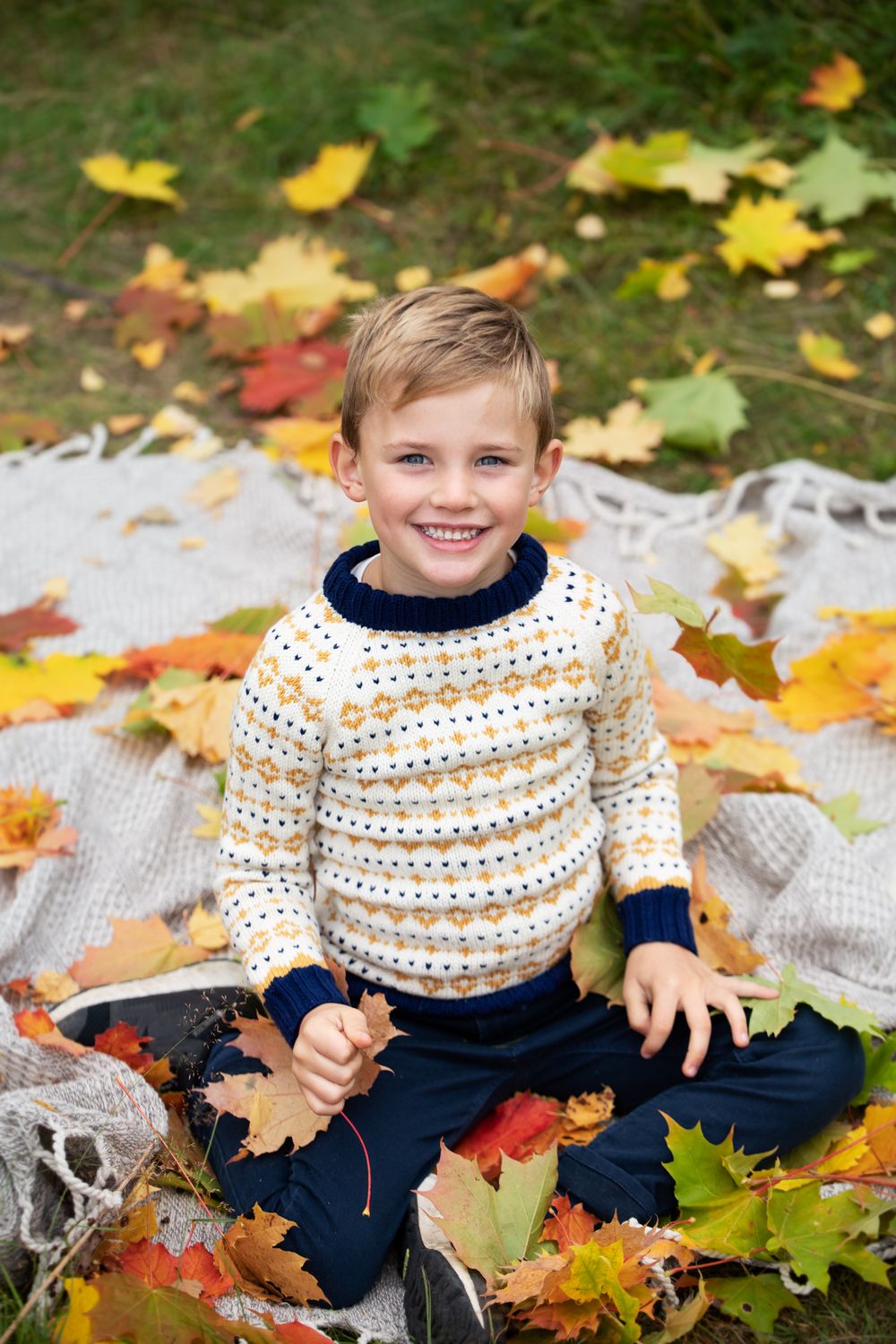 Smiling boy in patterned sweater sitting on blanket surrounded by colorful autumn leaves in a park.