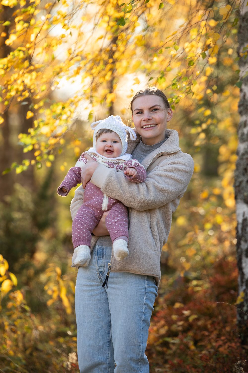 Smiling mother holding happy baby in autumn forest with golden yellow leaves in background.