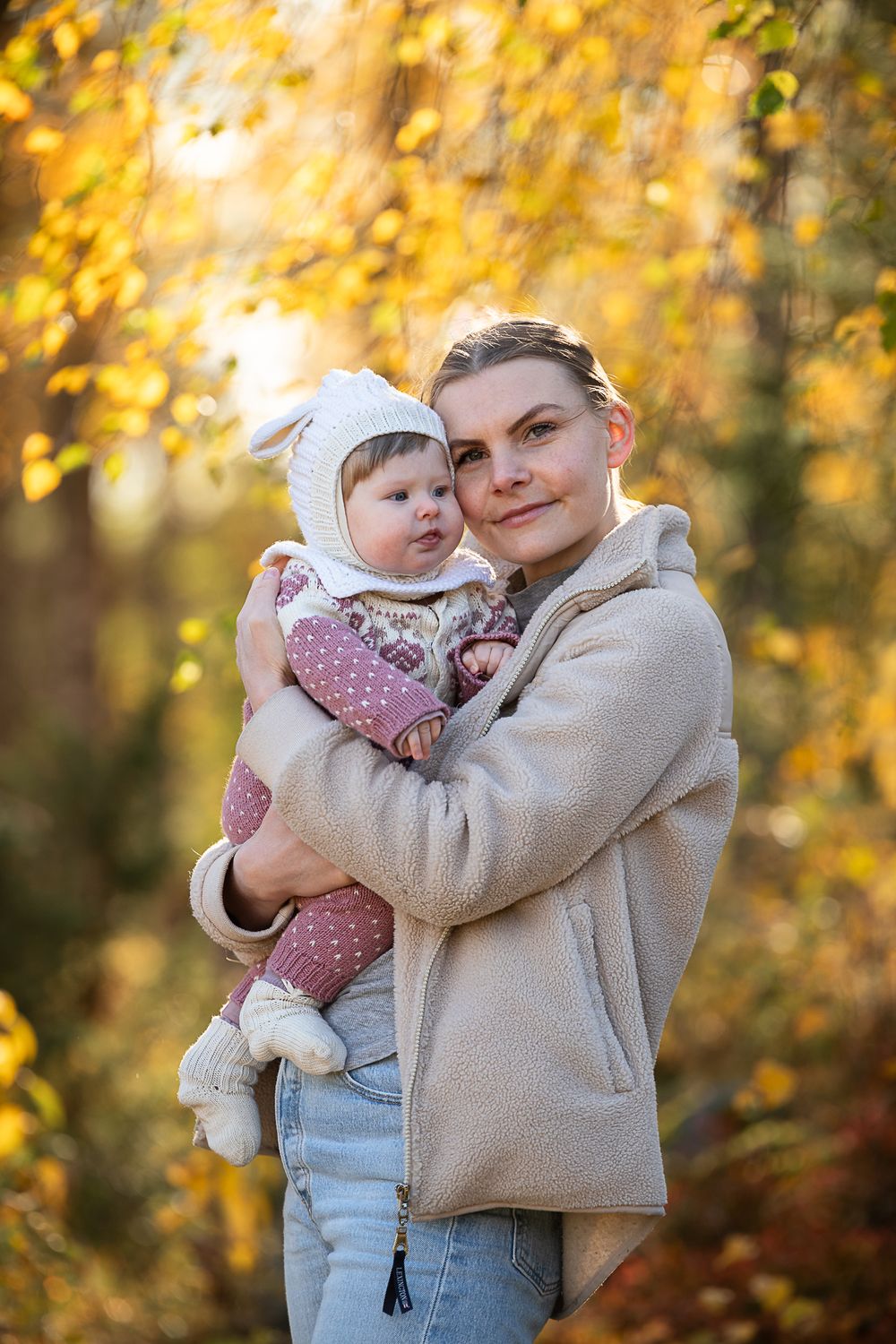 A young mother holds her baby in autumn forest, both wearing cozy outfits amid golden fall foliage.