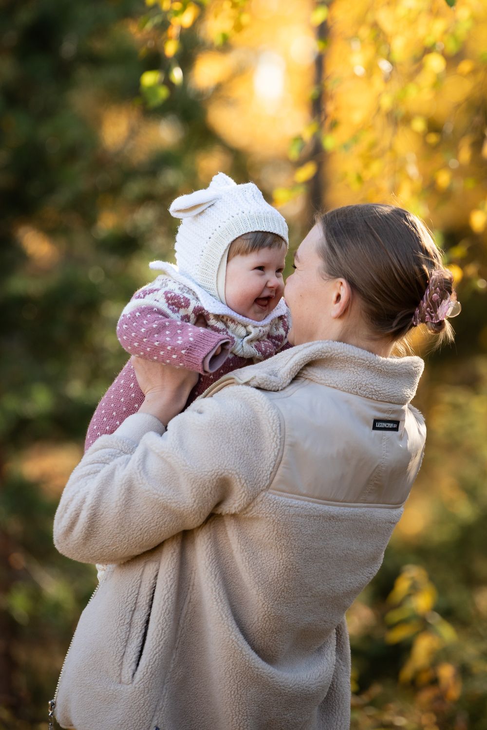 Mother holding smiling baby in white bunny hat outdoors in autumn forest with golden foliage background.