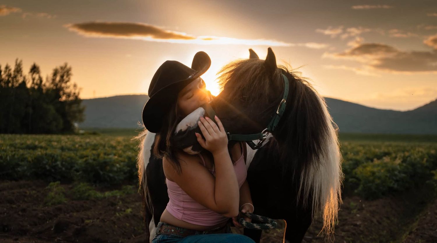 Jeune cavalière au chapeau de cowboy qui embrasse son cheval au coucher du soleil, devant les montagnes, lors d'une séance photo équestre.