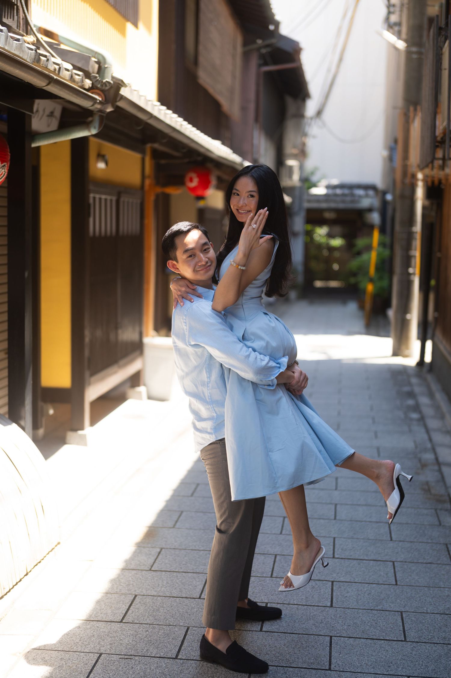 Smiling couple in coordinated blue outfits embracing as a man lifts a woman in a heeled sandals on a sunlit traditional stone-paved alley.