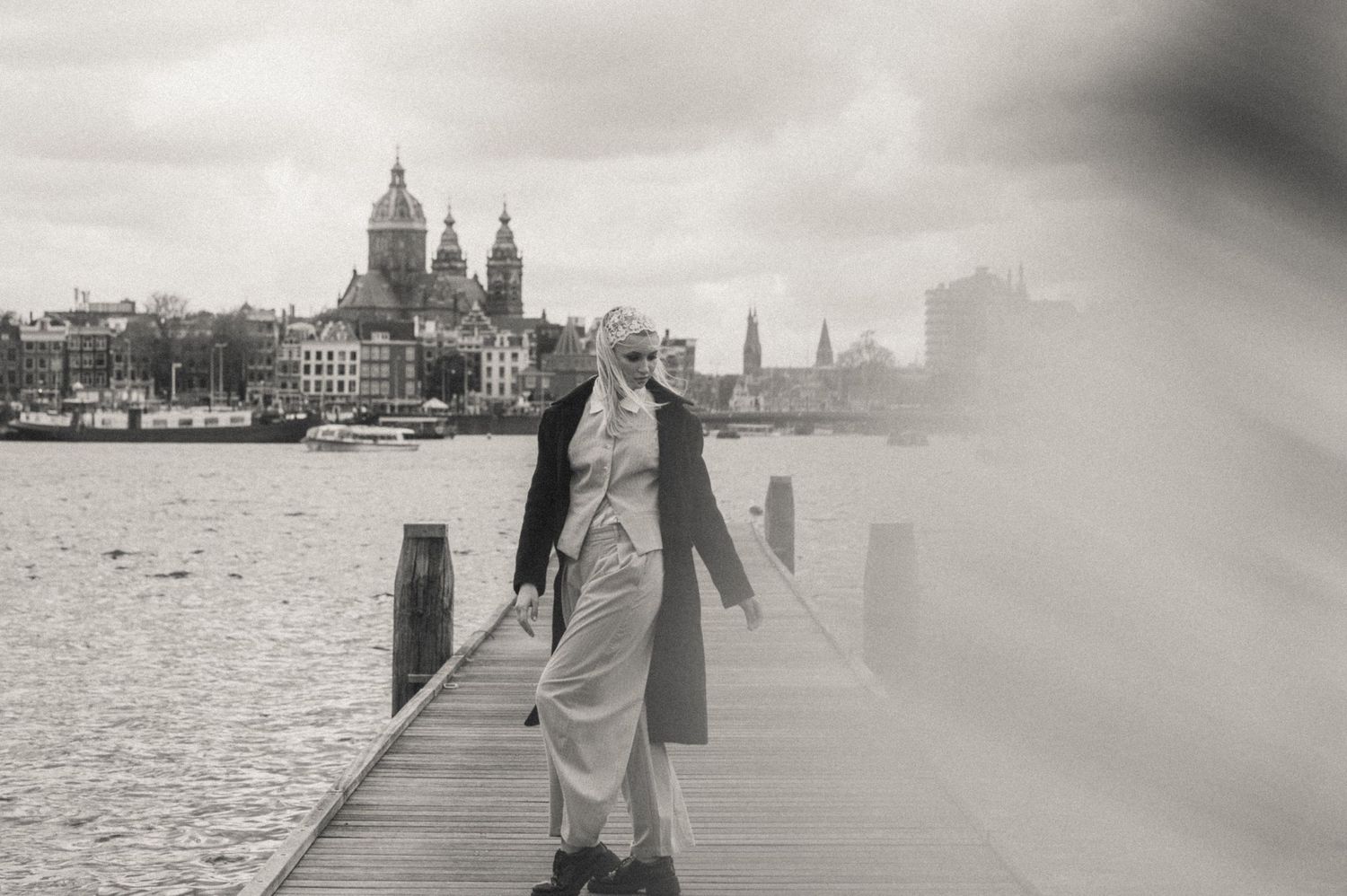 Woman walks along a wooden pier by calm water with a historic domed church and city skyline in black and white. Amsterdam