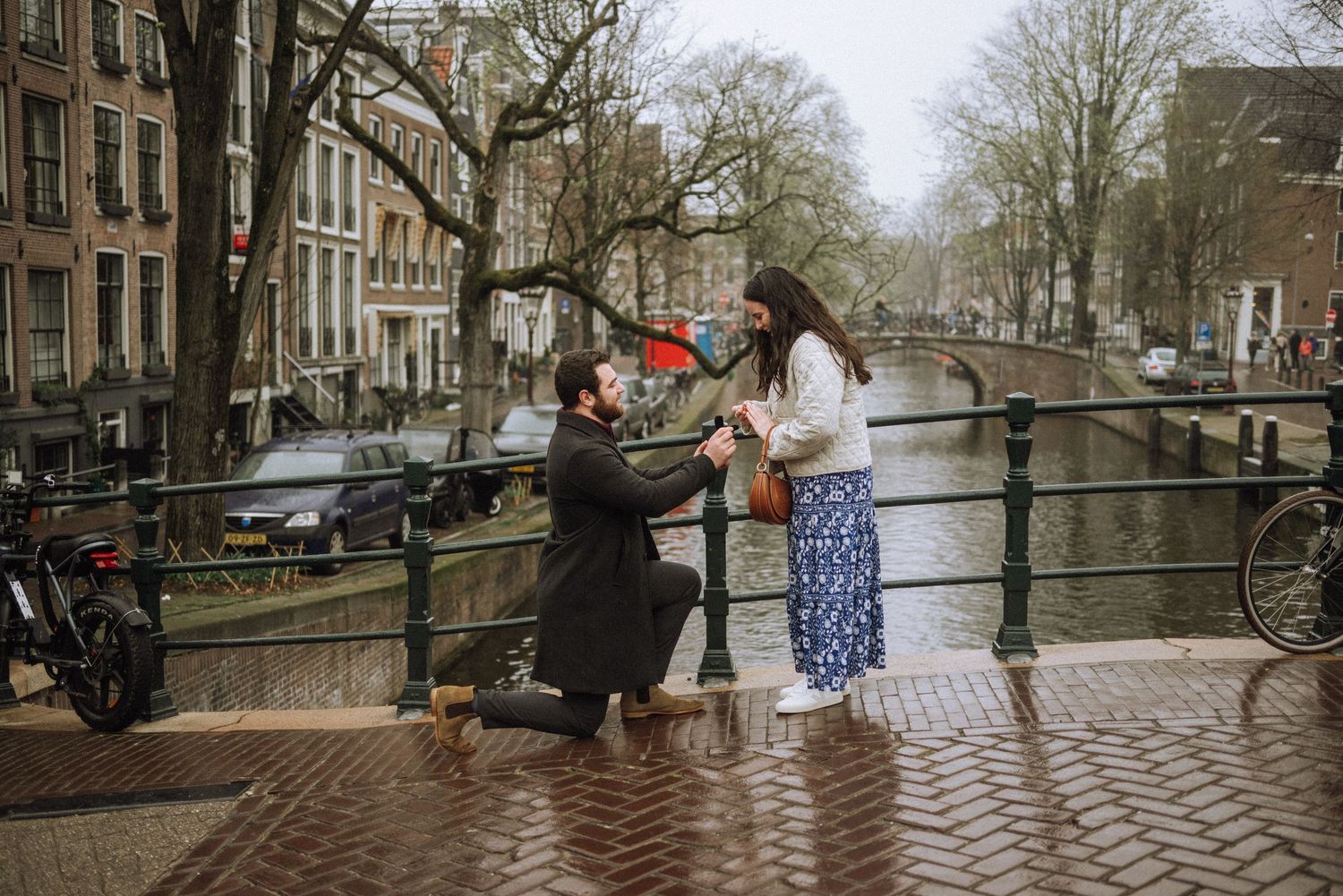 Man in a dark coat kneels on one knee proposing to a smiling woman on a brick canal bridge lined with trees and historic buildings.