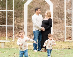 Pregnant woman in black dress with husband and two young boys in matching bear sweaters during outdoor maternity photo session.
