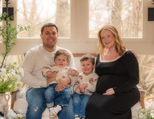 Smiling family of four with pregnant mother in black dress and father holding two toddlers on a couch.