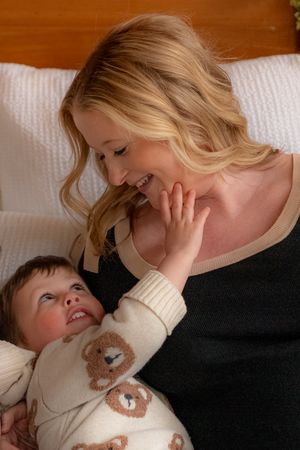 A smiling blonde woman holds a baby in a bear-print onesie, sharing a joyful moment together on a white couch.