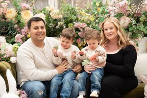 Family of four poses with colorful spring flowers, parents holding two young boys on a floral decorated bench.