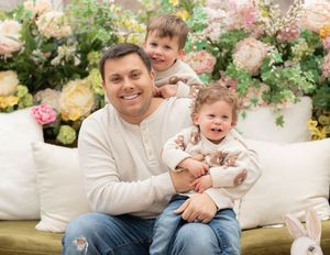 A smiling man holds two young children on a floral-decorated bench in a bright spring-themed photo setting.
