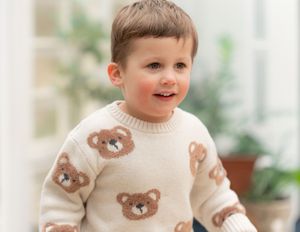 Toddler boy wearing a cream knit sweater with brown teddy bear pattern, smiling in a bright indoor setting.
