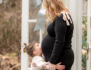Pregnant woman in black dress with toddler boy touching her baby bump by a decorated glass door.