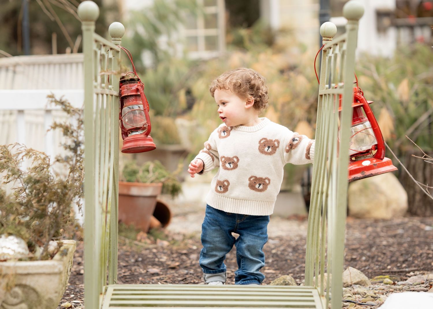 Toddler in bear sweater and jeans stands on garden bridge flanked by red lanterns.