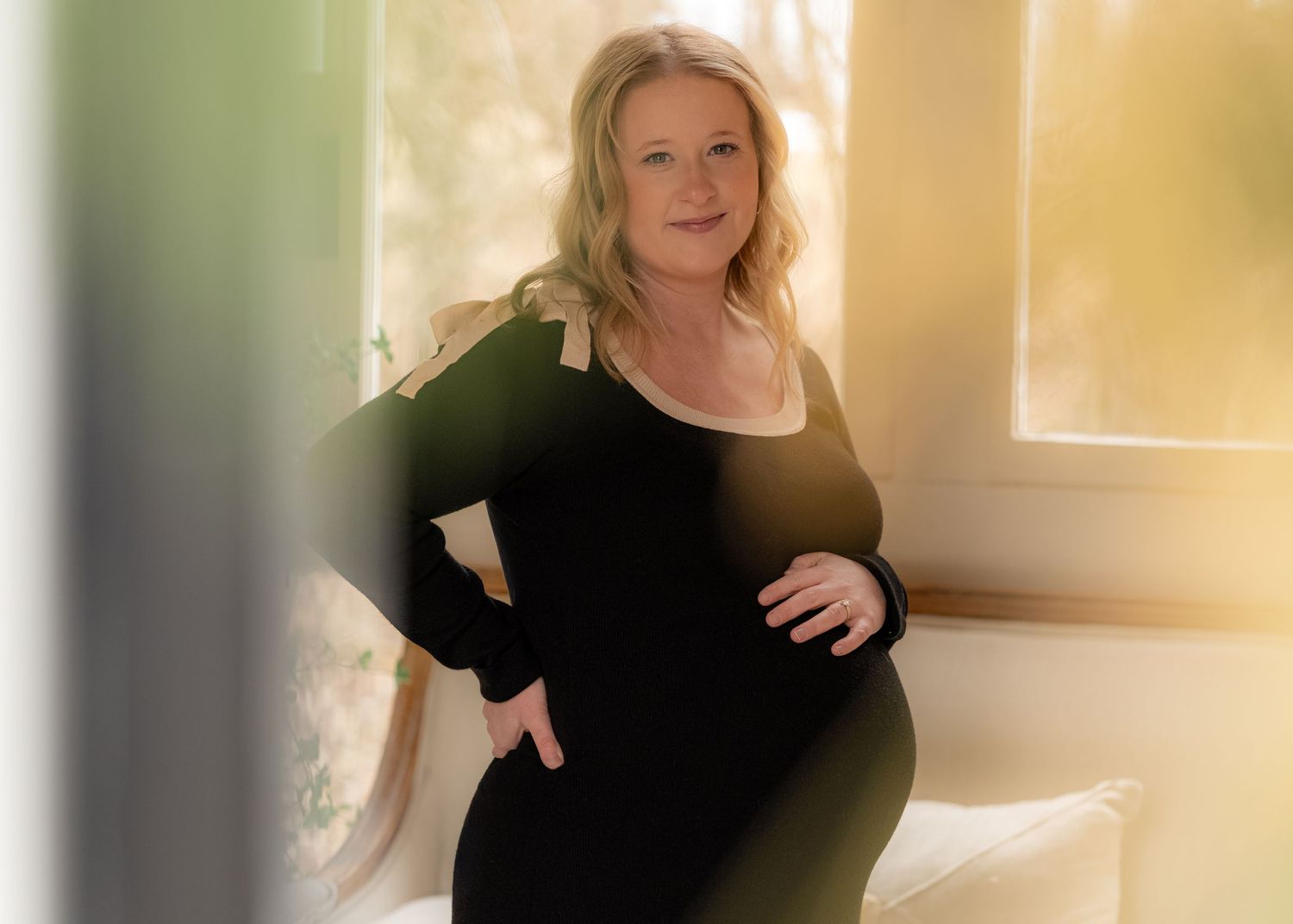 Pregnant woman in black dress posing by window with golden light.