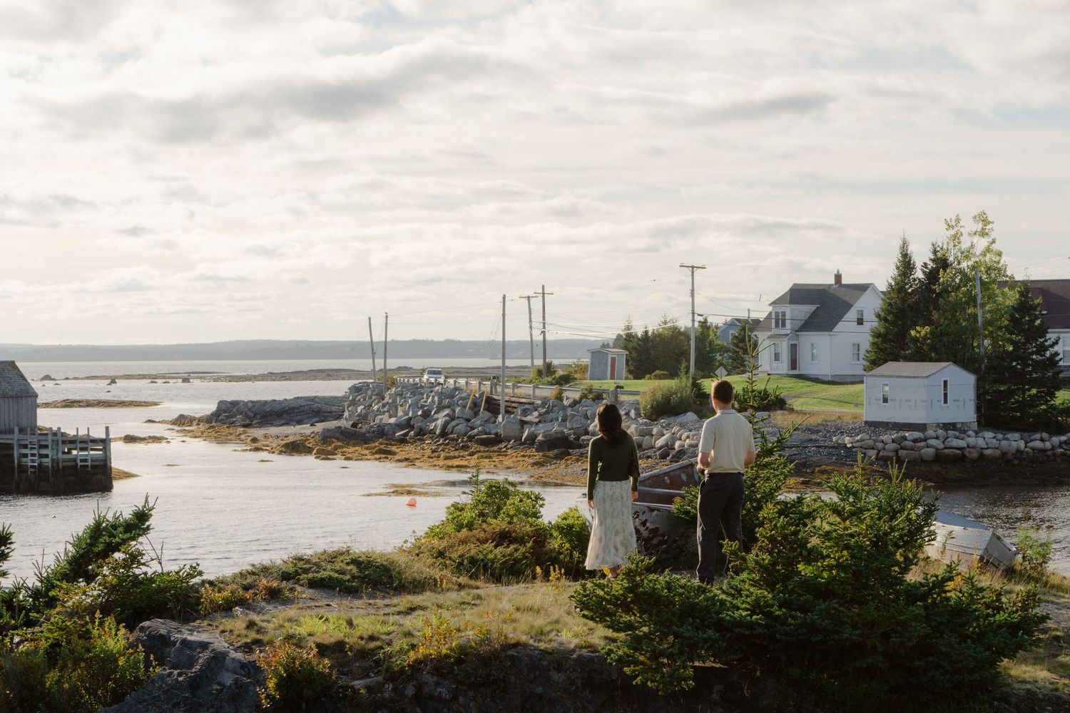 Two people stand overlooking a scenic coastal village with rocky shores, a white house, and utility poles under a cloudy sky.