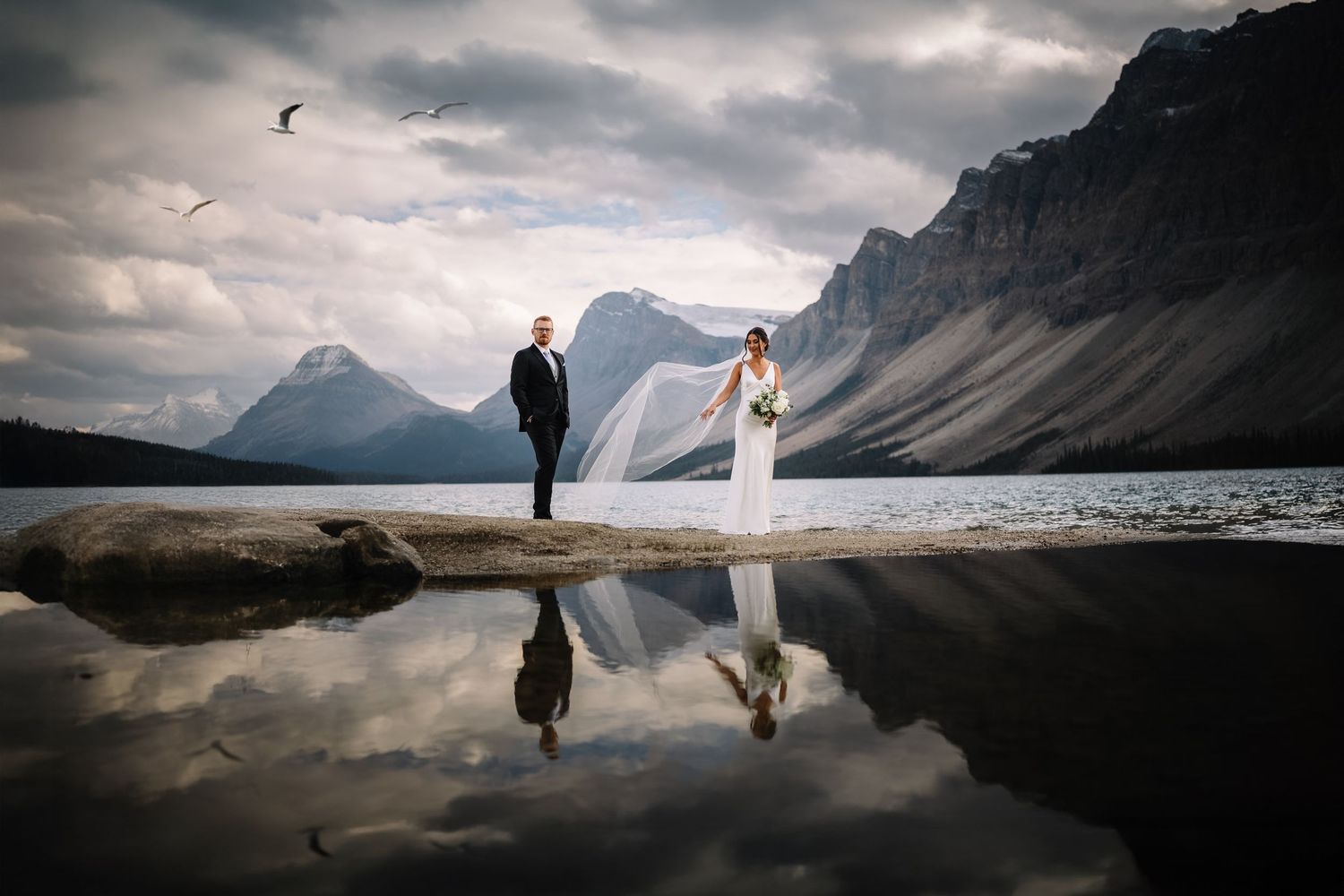 Bride and groom at a glassy mountain lake below rugged peaks, her veil in the wind, by a Banff wedding photographer