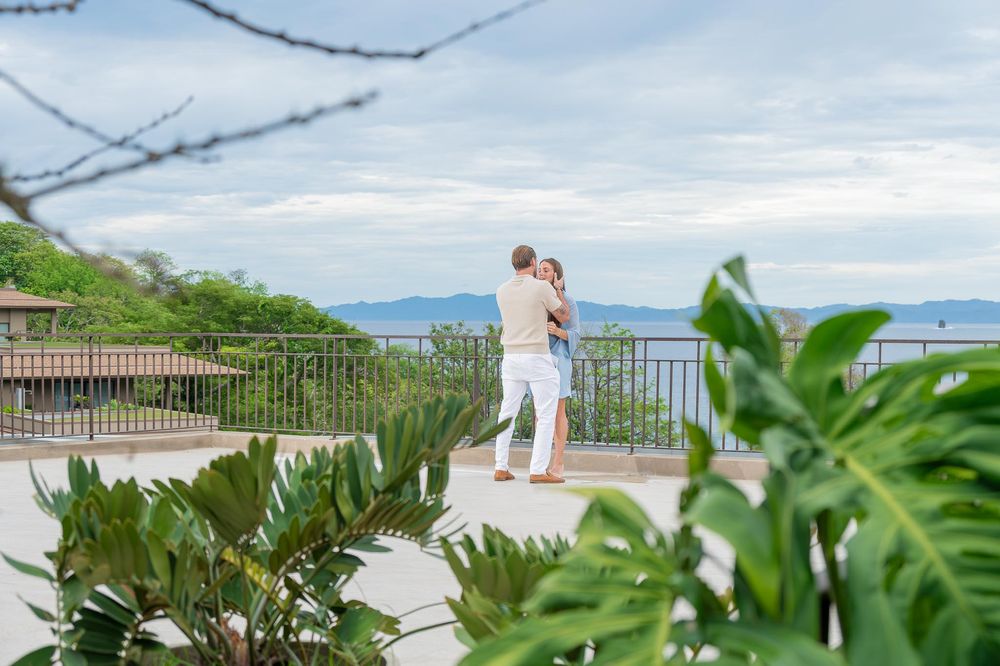 Couple embracing on a scenic overlook surrounded by tropical greenery with ocean views in the background.