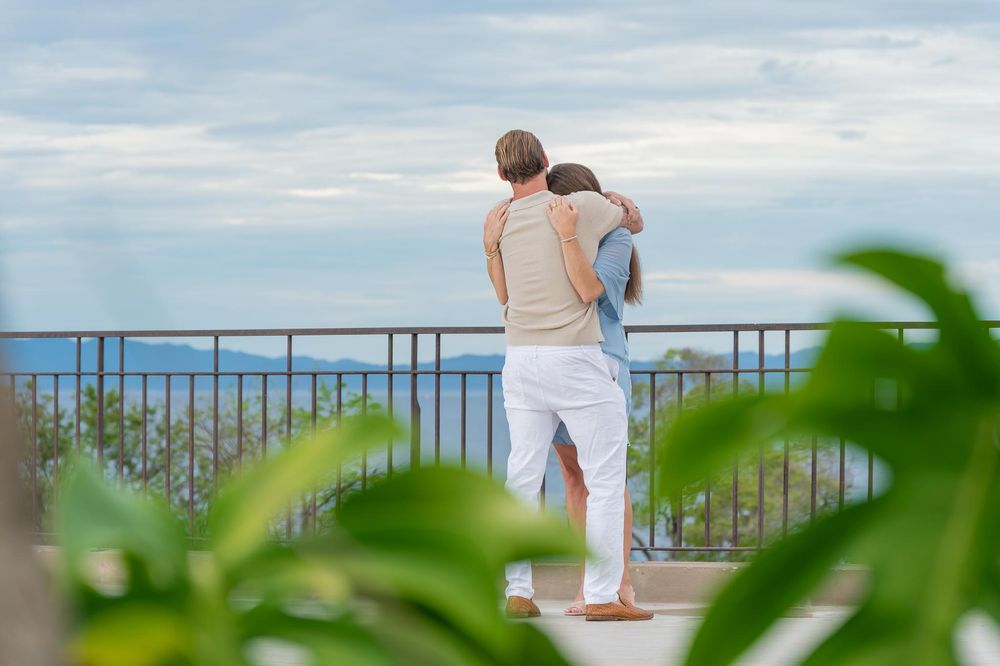 Couple embracing on a tropical balcony overlooking the ocean, surrounded by lush green foliage.
