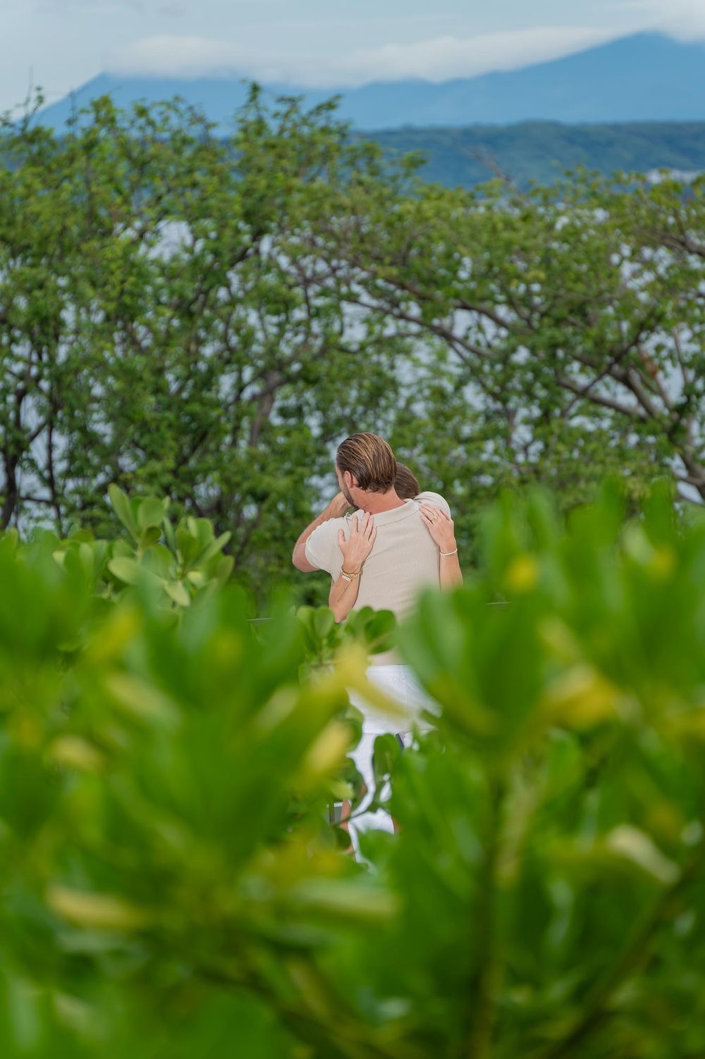 A woman stands among lush green tropical bushes with mountains visible in the background.
