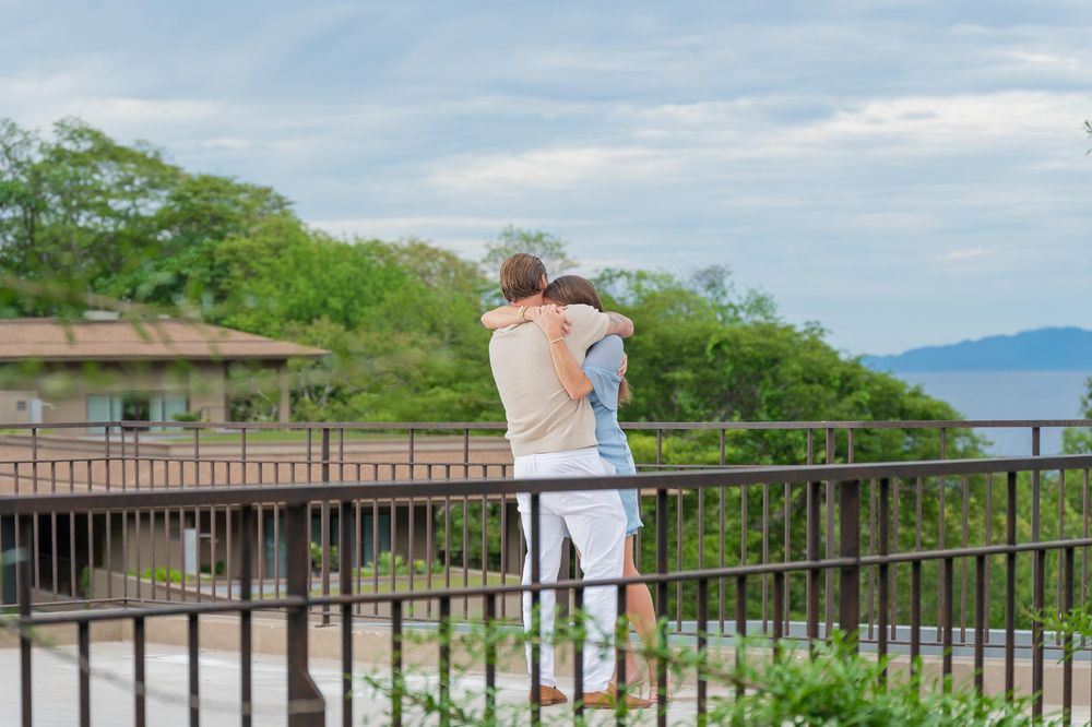 Couple embracing on a scenic overlook with lush green trees and calm water in the background.