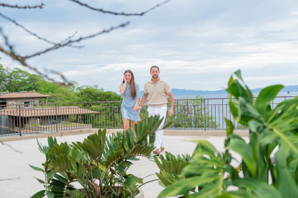 A couple walks together on a scenic terrace overlooking the ocean, surrounded by lush tropical greenery.